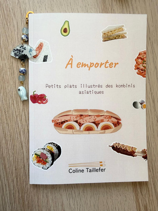 Un livre posé sur une table en bois, sa couverture illustrée de plats asiatiques colorés. Un marque‑page perlé, orné de petits charms, glisse depuis le haut. Le titre “À emporter” et le nom Coline Taillefer apparaissent en douceur.